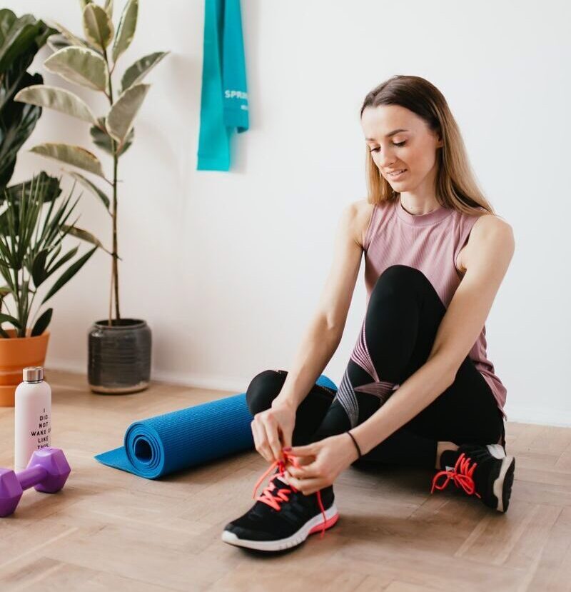 Full body young female athlete in sportswear tying shoelaces while sitting on floor near sport equipment and potted plants and looking down with smile