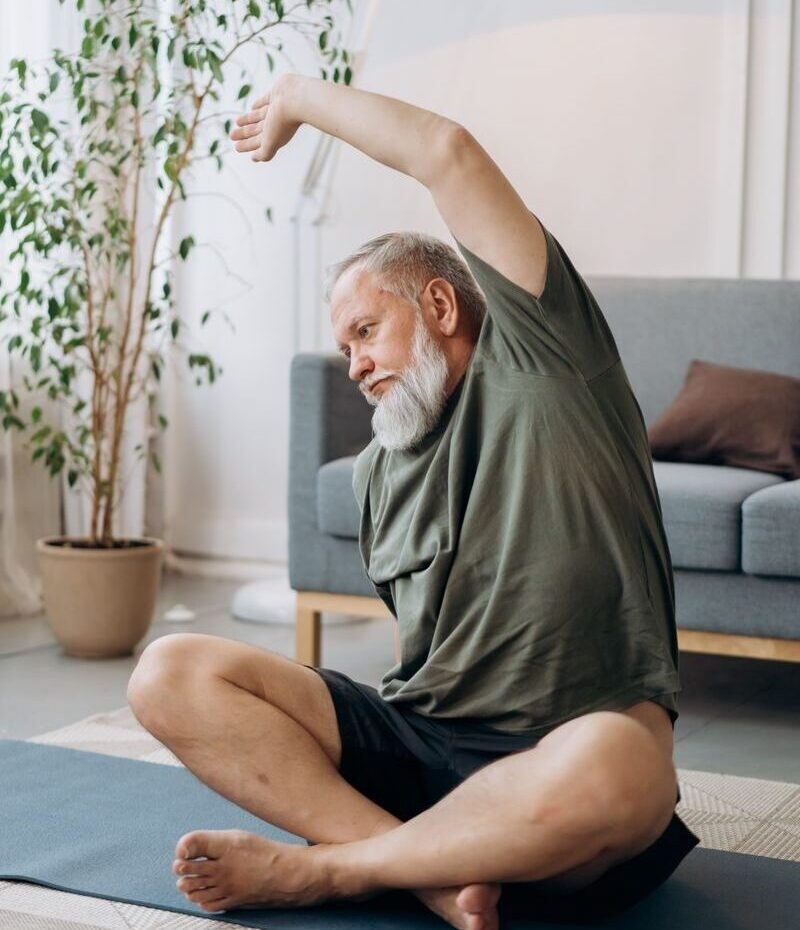 Elderly man stretching on a yoga mat in a cozy living room setting for wellness.